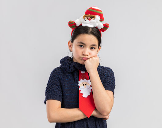 无聊小女孩穿着针织连衣裙 头上系着一条红色领带 戴着一个有趣的圣诞戒指 看上去又累又无聊新站着红色