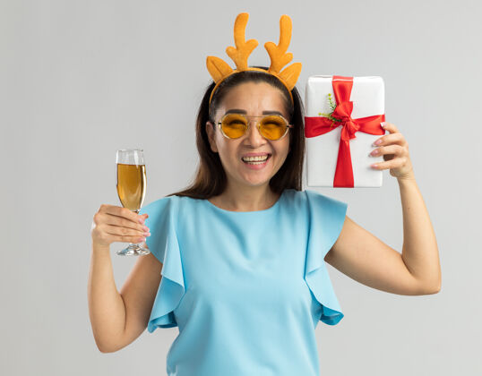 鹿身着蓝色上衣的快乐年轻女子 戴着滑稽的鹿角边和黄色眼镜 手里拿着一杯香槟和圣诞礼物 笑容满面蓝色香槟礼物