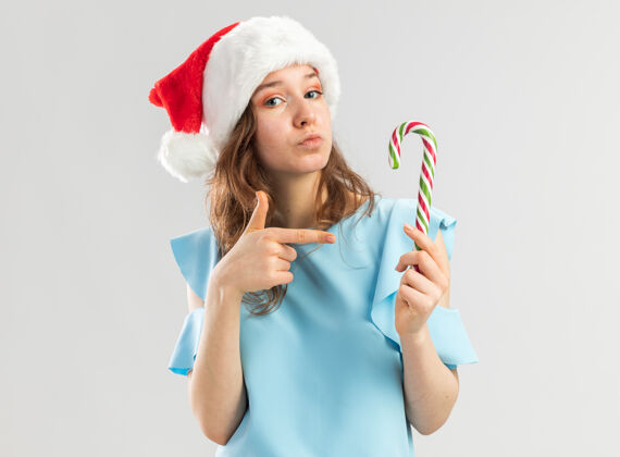 圣诞快乐身穿蓝色上衣 戴圣诞帽的年轻女子手持糖果手杖 用食指指着它 看上去很自信女人手指手杖