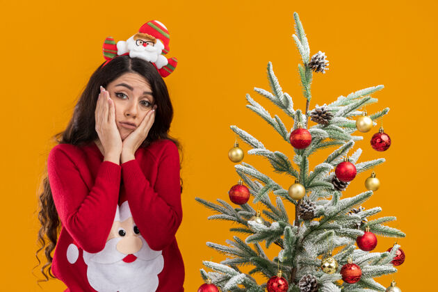 女孩戴着圣诞老人头巾和毛衣的悲伤的年轻漂亮女孩圣诞老人看圣诞树