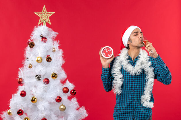 小伙子戴着圣诞老人帽 喝着一杯葡萄酒 拿着红色圣诞树旁的时钟 微笑着的年轻人喝酒年轻人站着