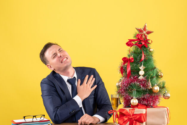 新郎前视图郁闷的男子抱着胸口痛苦地坐在圣诞树旁的桌子上 黄色的礼物坐着抱着商人