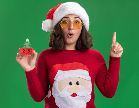 圣诞老人穿着圣诞毛衣的小女孩戴着圣诞帽 戴着眼镜 手里拿着25号玩具立方体 惊讶地伸出食指 站在绿色的墙上 有了新的想法圣诞节显示五
