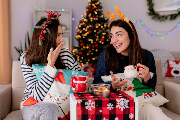 杯子笑容可掬的年轻姑娘们捧着杯子 看着彼此坐在扶手椅上 在家里享受圣诞节时光享受漂亮年轻