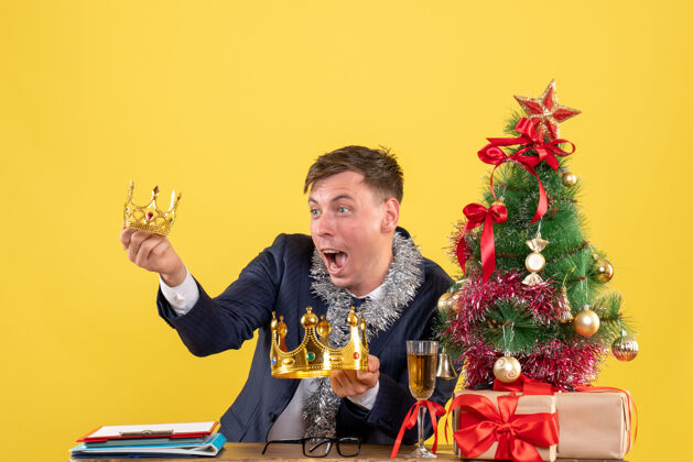 坐前视图的商人看着皇冠坐在圣诞树附近的桌子上 黄色的礼物人东方人人