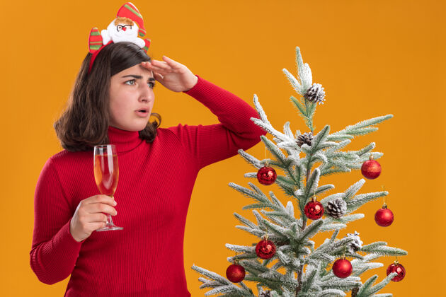 杯子穿着圣诞毛衣的年轻女孩戴着滑稽的头带 手里拿着一杯香槟 手举着头站在橙色墙上的圣诞树旁遥望远方香槟头带远