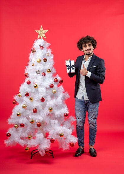 礼物年轻人在红色装饰的白色圣诞树旁展示他的礼物制作ok手势庆祝新年镜头圣诞节男人
