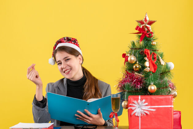 快乐前视图微笑的年轻女孩戴着圣诞帽坐在圣诞树和礼物鸡尾酒桌旁年轻女孩礼物桌子