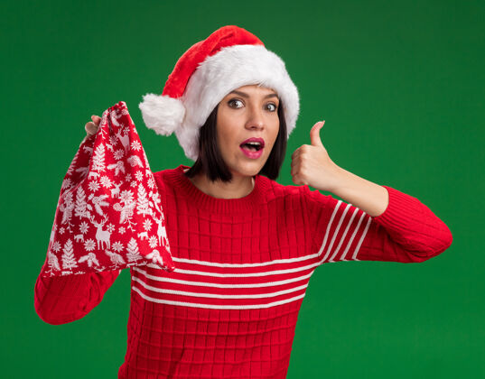 圣诞节令人印象深刻的年轻女孩戴着圣诞帽拿着圣诞礼物袋看着相机显示拇指向上孤立的绿色背景显示印象深刻袋子