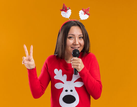 女人笑容满面的年轻亚洲女孩戴着圣诞发箍 穿着毛衣 在麦克风上讲话 在橙色的墙上显示出与世隔绝的和平姿态手势展示毛衣
