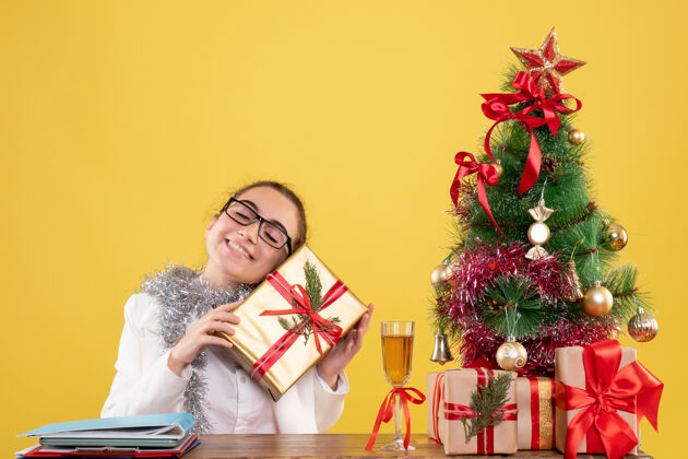 丝带前视图：女医生坐在桌子后面 拿着圣诞礼物和黄色背景上的树健康背景女医生