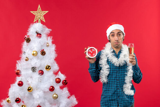 时钟一个戴着圣诞老人帽子 拿着一杯酒 站着闹钟的悲伤的年轻人礼物站着圣诞树