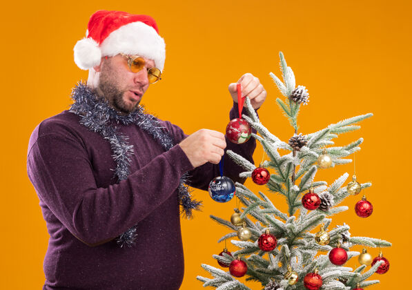 近困惑的中年男子戴着圣诞帽 脖子上戴着金箔花环 戴着眼镜站在圣诞树旁的侧视图中 用橙色背景上孤立的圣诞饰品装饰圣诞树花环穿着圣诞