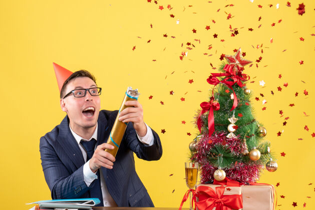 树前视图的商人与部分帽使用党波普尔站在桌子后面靠近圣诞树和黄色的礼物男人庆祝生意人