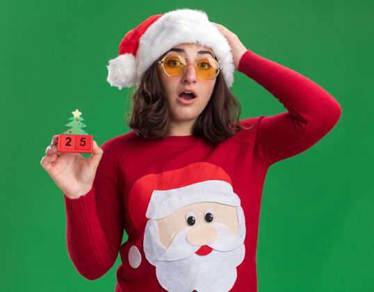帽子穿着圣诞毛衣的小女孩戴着圣诞帽 戴着眼镜 手里拿着25号的玩具立方体 站在绿色的墙上 惊讶不已五数字圣诞老人