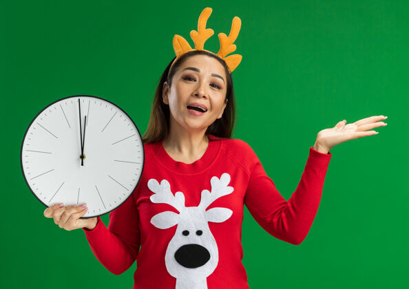 礼物快乐的年轻女子戴着鹿角和红色毛衣的圣诞戒指拿着挂钟看着相机微笑着用手臂站在绿色背景上呈现毛衣钟边