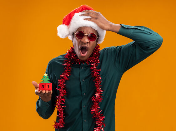 帽子戴着圣诞帽 戴着花环 戴着太阳镜的非裔美国人站在橘红色的墙上 手里拿着新年约会玩具方块 满脸愤怒 表情咄咄逼人立方体表情圣诞节