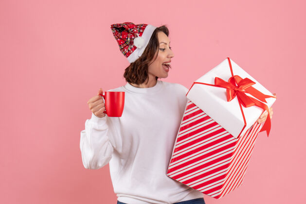 人年轻女子手持圣诞礼物和粉红色墙上的一杯茶的前视图圣诞节持有前面