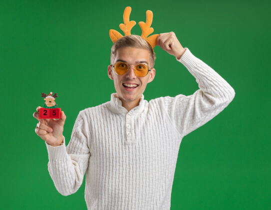 抓印象深刻的年轻帅哥戴着驯鹿鹿角头带戴着眼镜拿着圣诞树玩具抓日期头带隔离在绿色的墙上日期印象深刻头带