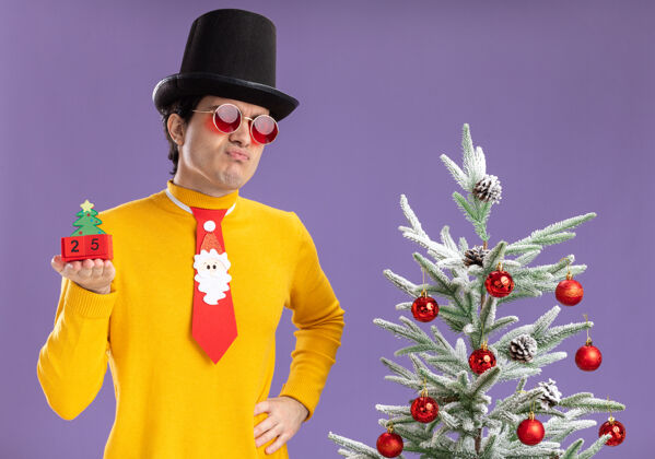 站着一个穿着黄色高领毛衣戴着眼镜的年轻人 戴着黑色帽子 打着滑稽的领带 手里拿着25号方块 上面写着日期日期圣诞树帽子