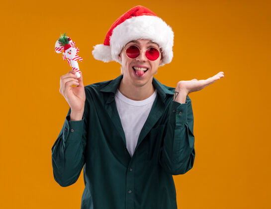 帽子戴着圣诞帽 戴着眼镜 拿着甜甜的藤条饰物 开玩笑的金发年轻人看着摄像机 在橙色背景上 他露出了舌头和空手装饰品手杖空