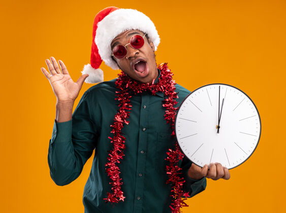 抱着戴着圣诞帽 戴着花环 戴着太阳镜 拿着时钟 惊讶地看着摄像机 抬起手臂站在橙色背景上站着惊讶举起来