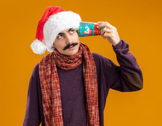 围巾一个留着胡子的男人戴着圣诞老人帽 脖子上围着暖和的围巾 耳朵上戴着彩色纸杯 站在橙色的背景下显得很困惑围着耳朵温暖