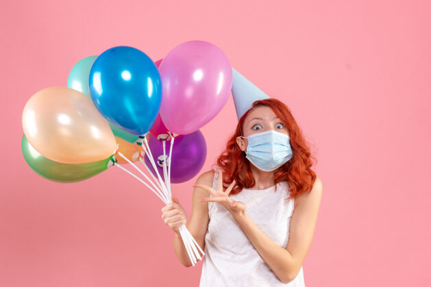 生日正面图是粉红色墙上戴着消毒面具手持彩色气球的年轻女子粉色气球举行