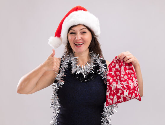 花环快乐的中年妇女戴着圣诞帽 脖子上戴着金箔花环 手里拿着圣诞礼物袋 看着相机 在白色背景上孤立地竖起大拇指快乐礼物围着