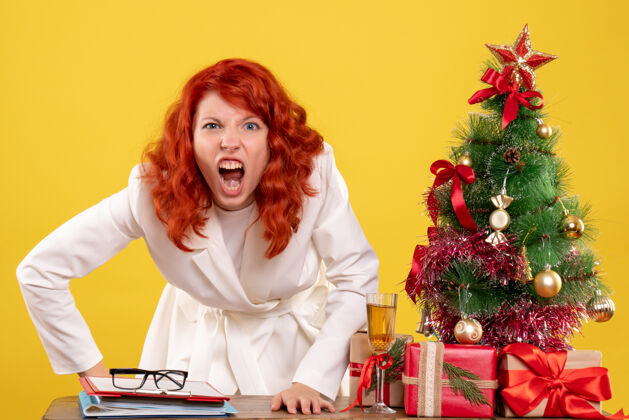 快乐正面图：女医生坐在桌子后面 拿着黄色背景上愤怒的圣诞礼物 还有圣诞树和礼盒礼物女医生圣诞节
