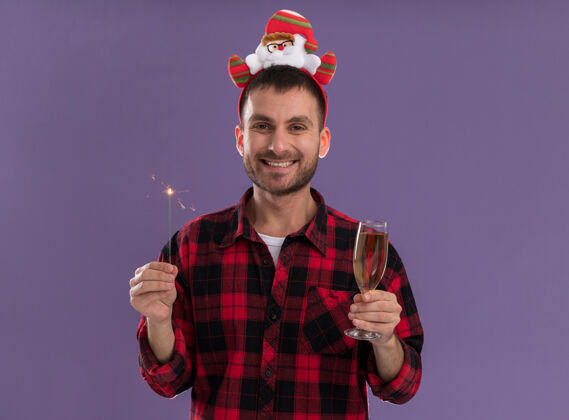 头带快乐的白人年轻人戴着圣诞老人的头带 手里拿着节日的火花灯和一杯香槟 隔离在紫色的墙上 留着复制空间年轻男人圣诞老人
