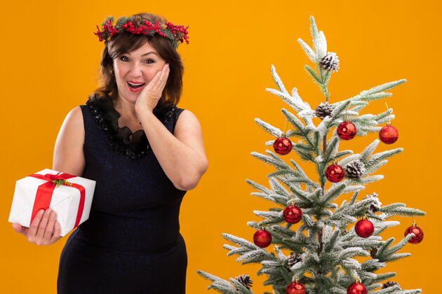 礼物兴奋的中年妇女头戴圣诞花环 脖子上戴着金属丝花环 站在装饰好的圣诞树旁 手里拿着礼包包头脸