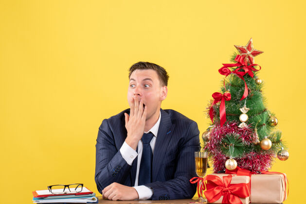 人前视图惊讶的人坐在圣诞树附近的桌子上 并提出了黄色墙壁上的自由空间商务人办公室