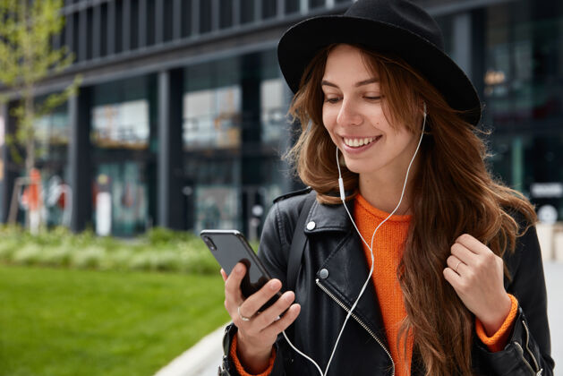 通信户外拍摄的高加索女性喜欢听音轨 使用现代手机和耳机 在市中心摆姿势 有着牙齿般的笑容技术应用程序博客