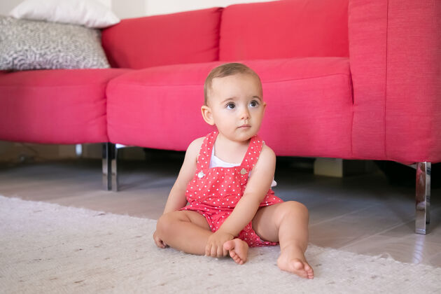 生活可爱的小宝宝光着脚坐在客厅的地毯上穿着红色工装裤的有趣沉思的女孩看着某人 抚摸着她的腿周末 童年和在家的概念婴儿期可爱童年