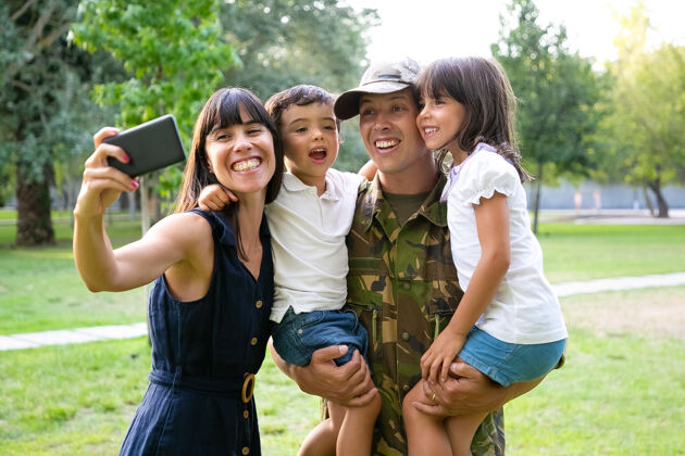 摆姿势快乐兴奋的军人 他的妻子和两个孩子庆祝爸爸回来 在公园里享受休闲时光 在手机上自拍中镜头家庭团聚或回家的概念女儿电话拍摄