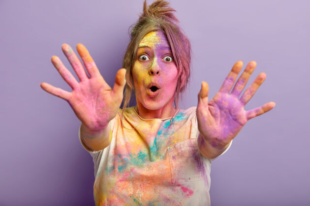 白种人照片中 一个惊讶有趣的女人 脸 手掌和衣服都是彩色的 庆祝胡里节 玩弄颜色 伸手 在紫色的墙上用粉末染料隔离节日玩耍粉末