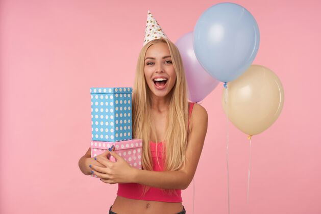 无包装身着粉色上衣 头戴节日帽 满头长发 喜气洋洋 充满魅力的年轻女子 在粉色背景下 在五颜六色的气球里摆出各种姿势 为得到生日礼物而感到兴奋和惊讶盒子礼物室内