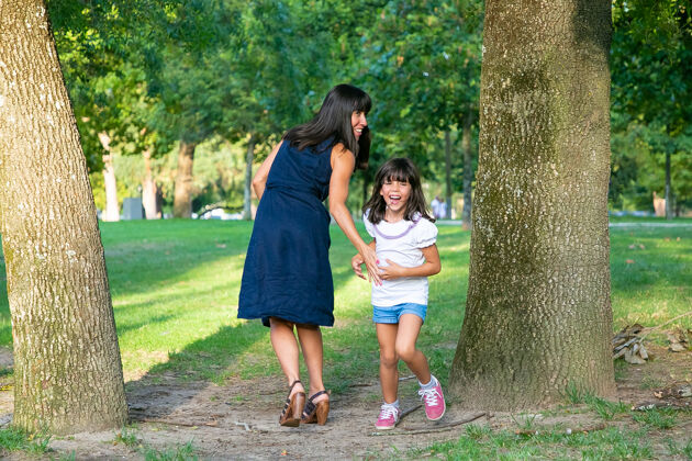 活跃兴奋快乐的女孩在户外和妈妈一起玩游戏 站在公园的树边笑全长家庭户外活动和休闲概念长度黑色头发女儿