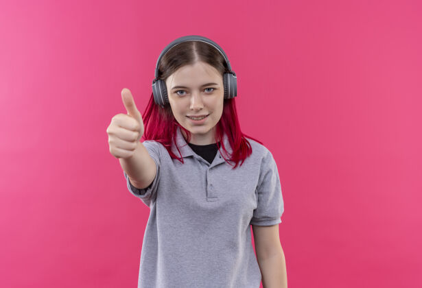复制微笑着的年轻漂亮女孩戴着灰色t恤 戴着耳机 大拇指竖立在孤立的粉色背景上 留有复印空间拇指女孩T恤