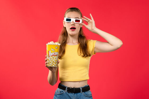 风景正面图年轻女性在电影院拿着爆米花包戴着d太阳镜在红墙电影院看女性趣味电影爆米花电影年轻的女的