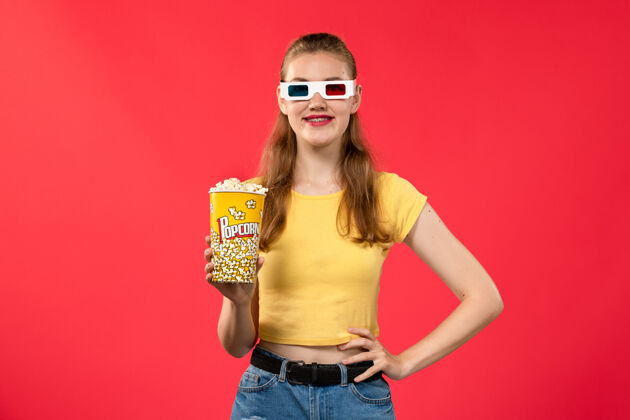 剧院正面图年轻女性在电影院拿着爆米花包戴着d太阳镜在红墙电影院看女性趣味电影漂亮的成人的年轻的