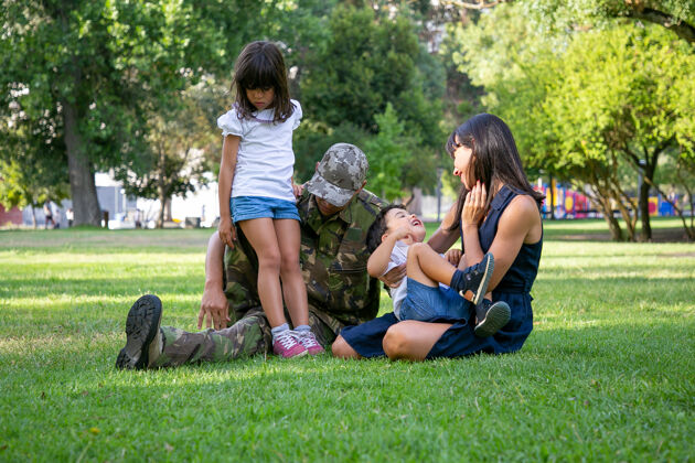 孩子快乐的一家人坐在城市公园的草地上身着军装的白人中年父亲 微笑着的母亲和孩子们一起在草地上放松家庭团聚 周末和回家的概念女人孩子军队