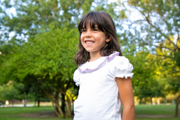 站立快乐的小黑发女孩站在城市公园里 看着远方 微笑着孩子们在夏天享受户外休闲时光中镜头童年的概念白种人活动小