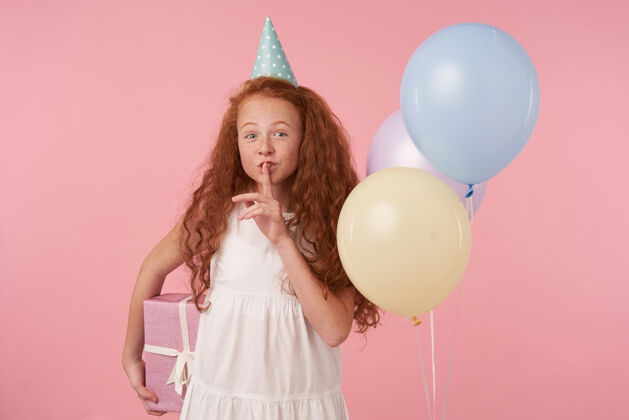 肖像积极的小红发孩子庆祝节日在粉红色的背景下穿着白色的裙子和生日帽拿着礼品盒 举手向她嘴在安静的姿态 要让惊喜童年惊喜严重