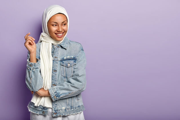 传统无忧无虑的深色皮肤的阿拉伯女模特开心地笑着 表达真挚的感情 笑容灿烂 聚精会神地放在一边 戴着头饰和牛仔夹克在紫色的墙上摆姿势 空白处满意自由高兴