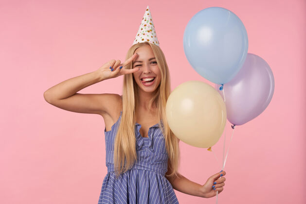 年轻有趣的室内镜头 年轻的金发女子长发眨眼 并提出了确定的姿态高兴 持有五彩气球在她的手上 而在粉红色的背景摆姿势圆锥形气球庆祝