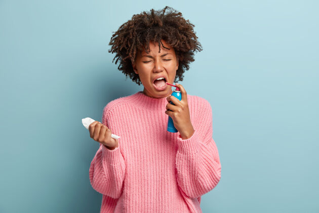 支气管炎绝望的女性患有咽喉疼痛 感觉不健康和不适 喷雾剂溅到嘴里 拿着纸巾 穿着超大套头衫 隔离在蓝色墙壁上治疗流感或感冒不满喷溅肖像