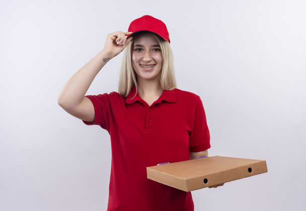 披萨微笑着送货的年轻女孩穿着红色t恤 戴着帽子 戴着牙套 手里拿着披萨盒 把她的手放在隔离的白色背景上的帽子上送货白色抱着