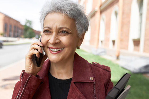 电话穿着夹克的中年妇女的头像在城市街道上摆着姿势 耳朵边放着手机灰色电话成熟